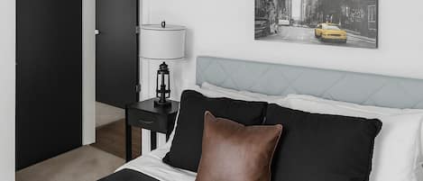 A bedroom with a bed, a nightstand, a lamp, a door, and a cityscape painting on the wall.