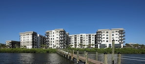 Ein Wohngebäude am Wasser mit mehreren Balkonen, einer Holzsteganlage und einem strahlend blauen Himmel.
