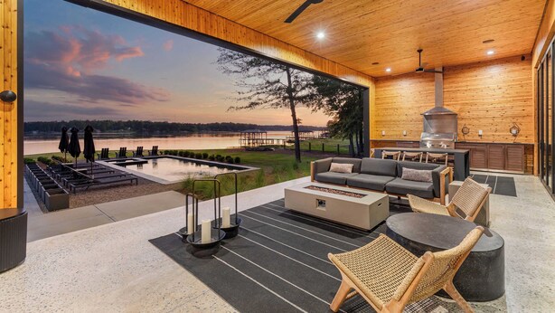 A modern outdoor living space with a large window overlooking a lake, a barbecue area, and a seating arrangement. - Dazzling Design Meets Spectacular Views (Eatonton)