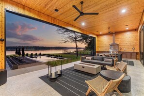 A modern outdoor living space with a large window overlooking a lake, a barbecue area, and a seating arrangement. - Dazzling Design Meets Spectacular Views (Eatonton)