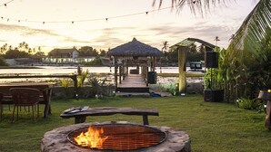 An outdoor patio with a stone fire pit, a thatched-roof gazebo, and a view of a body of water. - Hotel Boutique Quinta Três Marias (Paracuru)