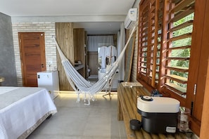 Una habitación con una cama, una hamaca, un banco de madera, una mesita y una ventana que da a zonas verdes.
