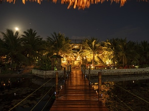 A wooden bridge leading to a building with a thatched roof, surrounded by palm trees and water. - Hotel Boutique Quinta Três Marias (Paracuru)
