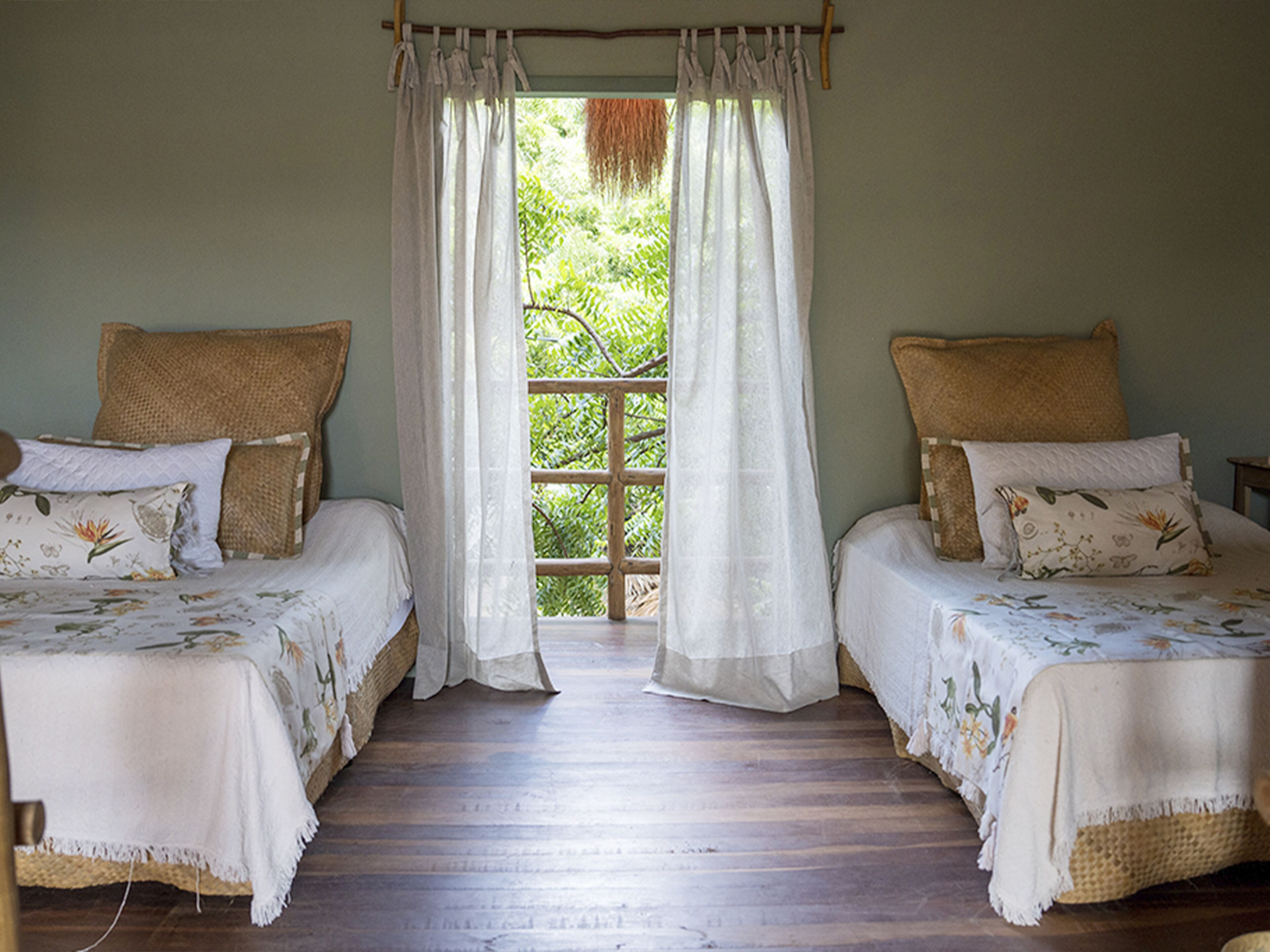 A room with two beds, each with a woven headboard, a window with white curtains, and a view of greenery outside.