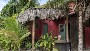 A thatched-roof structure with a red wall and green shutters, surrounded by tropical vegetation and a sandy ground. - Hotel Boutique Quinta Três Marias (Paracuru)