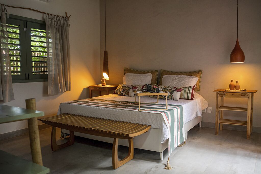 Un dormitorio con cama, mesitas de noche, un banco, una lámpara colgante y un ventanal con cortinas.