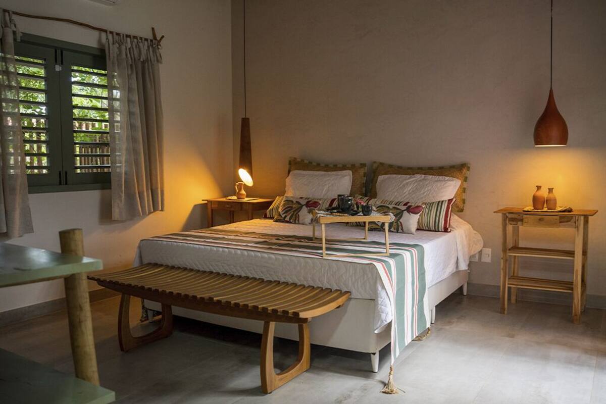 Un dormitorio con cama, mesitas de noche, un banco, una lámpara colgante y un ventanal con cortinas.