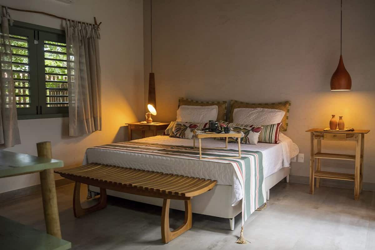 Un dormitorio con cama, mesitas de noche, un banco, una lámpara colgante y un ventanal con cortinas.