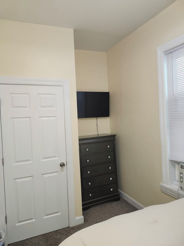 A bedroom with a white door, a black dresser, a flat-screen TV mounted above it, and a window with blinds.