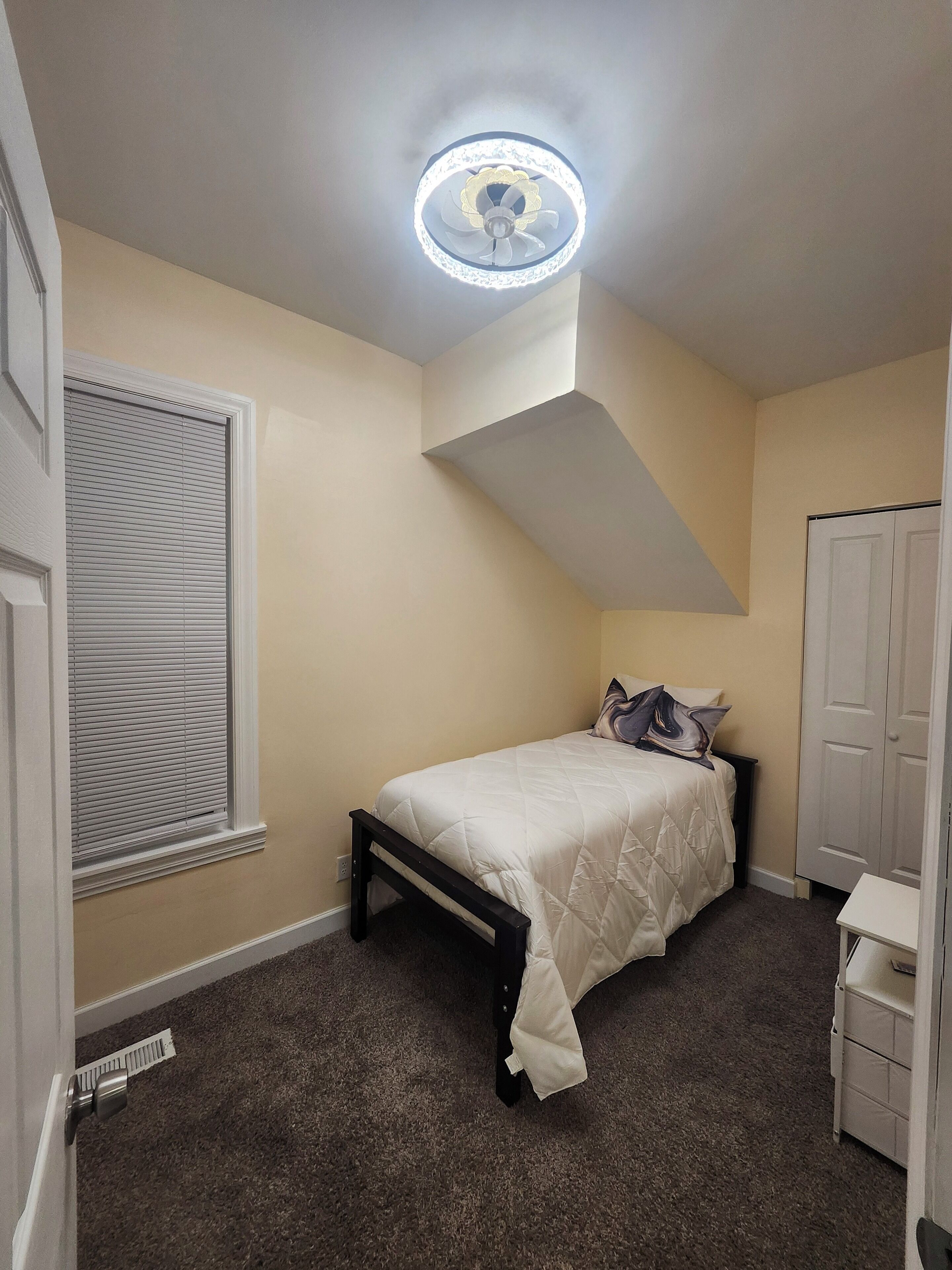A bedroom with a bed, a ceiling fan, a window with blinds, a wardrobe, and a carpeted floor.