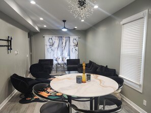 A dining area with a round table, black chairs, a chandelier, and a window with blinds.