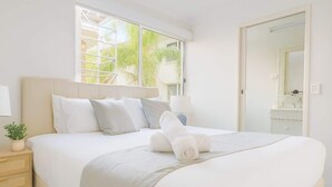 A neatly made bed with pillows and a rolled towel, a bedside table with a plant, a window with a view of palm trees, and a bathroom with a mirror and sink. - Broadbeach | 2Min to Beach | Pool | Free Parking (Broadbeach)