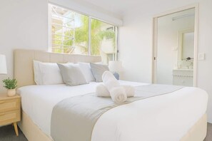 Ein ordentlich bezogenes Bett mit Kissen und einem zusammengerollten Handtuch, ein Nachttisch mit Pflanze, ein Fenster mit Blick auf Palmen und ein Badezimmer mit Spiegel und Waschbecken.