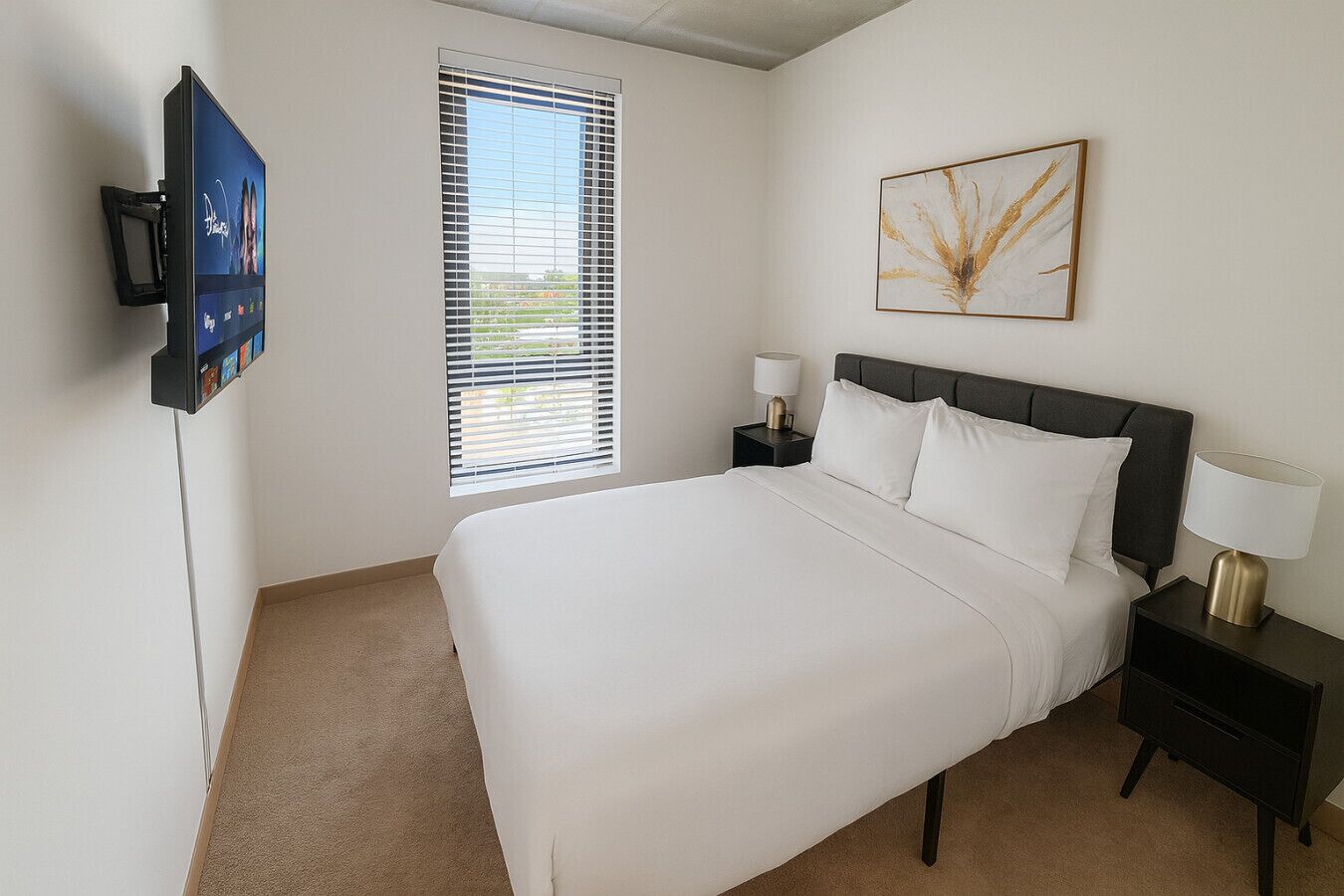 A hotel room with a bed, bedside tables, a TV, a window with blinds, and a wall-mounted artwork.
