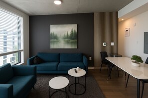 A modern living room with a dark-colored sofa, a coffee table, a dining table with chairs, a potted plant, and a painting of a forest on the wall. - Five Points South | Spacious 2BR w/ 2 Queens D5 (Birmingham)