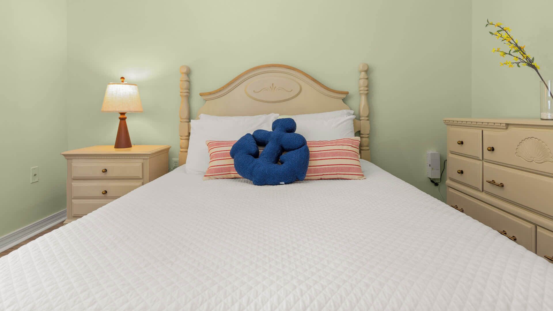 A bed with a headboard, a bedside table with a lamp, a dresser, and a vase with flowers.