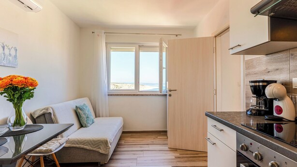 Een kleine keuken met een fornuis, koffiezetapparaat en uitzicht op het strand door een geopende deur.