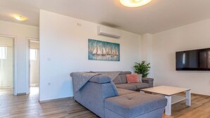 Ein modernes Wohnzimmer mit einem grauen Ecksofa, einem weißen Couchtisch, einem an der Wand montierten Fernseher und einem Gemälde mit Segelbooten.