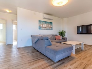 Ein modernes Wohnzimmer mit einem grauen Ecksofa, einem weißen Couchtisch, einem an der Wand montierten Fernseher und einem Gemälde mit Segelbooten.