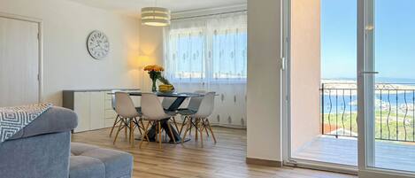 A modern living room with a sofa, dining table, chairs, and a view of the sea through a balcony.