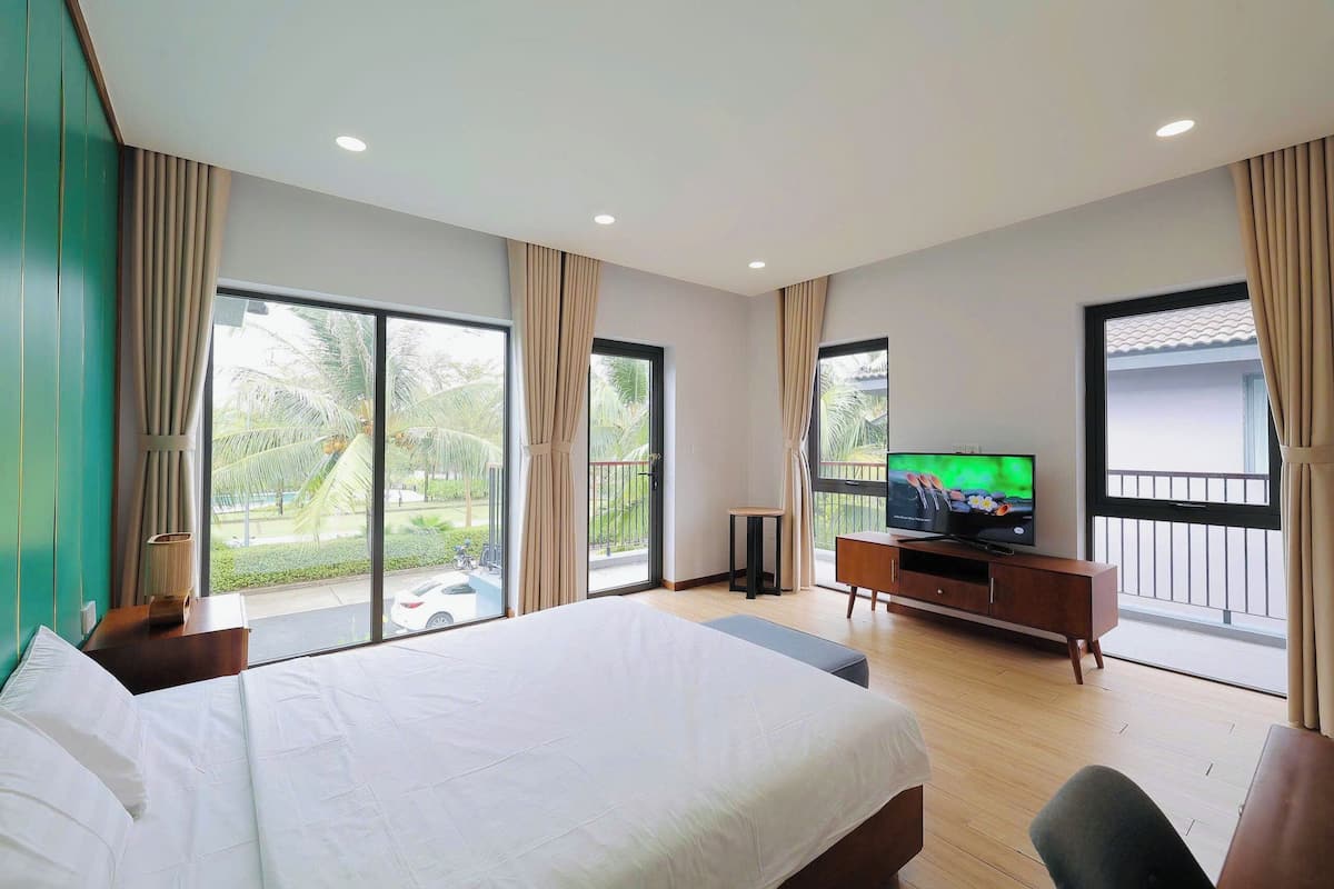 A hotel room with a large bed, a TV, a wooden cabinet, and a balcony with a view of palm trees.
