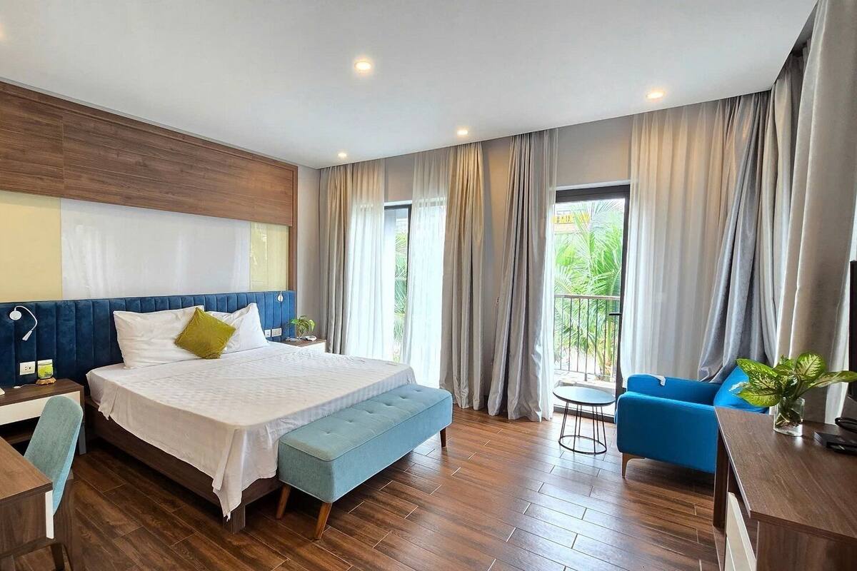A hotel room with a bed, a desk, a chair, a blue sofa, a TV, and a balcony with curtains.