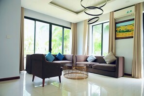 A modern living room with a sofa, armchair, and coffee table, large windows with curtains, and a painting on the wall. - V-Luxury Villas Phu Quoc (Phú Quôc)