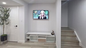 A modern living room with a flat-screen TV mounted on the wall, a white bench, a staircase, and a potted plant.