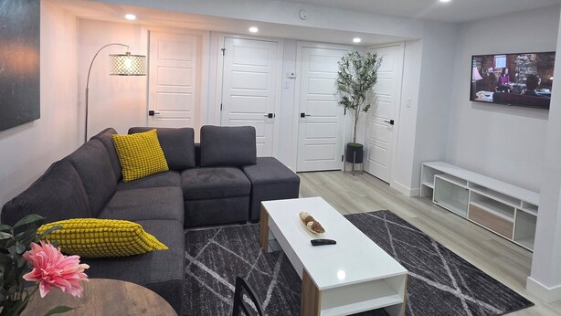 A modern living room with a sectional sofa, a coffee table, a dining table with chairs, recessed ceiling lights, a wall-mounted TV, and a potted plant.