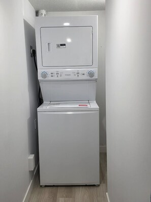 A compact, white laundry room with a front-loading washer and dryer, a small shelf above the dryer, and a wall-mounted electrical outlet. - Sleep in Silverstone queen size bed & enjoy our 7-Stage Water filtration System. (Edmonton)