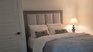 A bedroom with a bed, a nightstand with a lamp, a door, and a wooden floor.