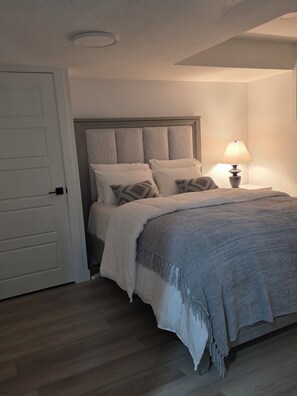 A bedroom with a bed, a nightstand with a lamp, a door, and a wooden floor. - Sleep in Silverstone queen size bed & enjoy our 7-Stage Water filtration System. (Edmonton)
