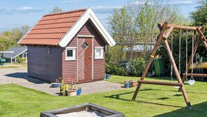 A small wooden house with a red roof, a swing set, and a sandbox in a grassy yard. - Charming holiday home near the beach (Hejls)