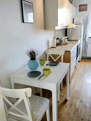 Cocina compacta con muebles blancos, una mesa de comedor con sillas y una pequeña planta en maceta.