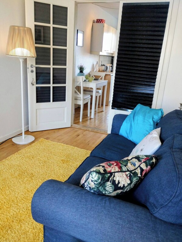 A living room with a sofa, a yellow rug, a lamp, and a kitchen area in the background.