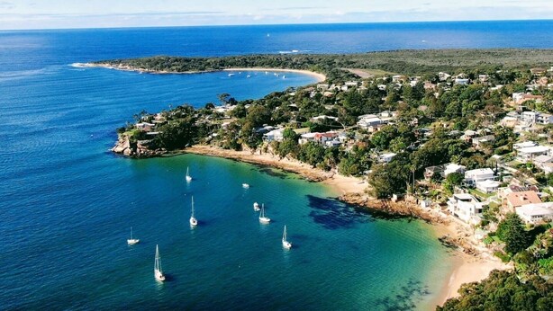Vue aérienne d’une ville côtière, avec des maisons le long du littoral, une mer bleue et limpide, et plusieurs voiliers ancrés près du rivage.
