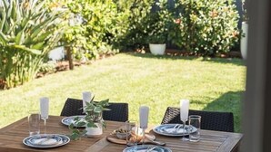 Un ensemble de mobilier de patio en bois, comprenant une table pour six personnes et des chaises en osier, entouré de plantes en pot et d’une clôture.