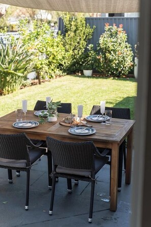 A wooden patio table set for six with wicker chairs, surrounded by potted plants and a fence. - Charming Sans Souci Cottage with Garden & Verandah (Sans Souci)