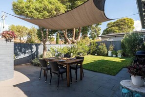 A patio with a table set for four, surrounded by chairs, under a shade sail.