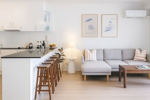 Eine moderne Küche mit weißen Schränken, einer schwarzen Arbeitsplatte und Holzstühlen. Ein graues Sofa mit gestreiftem Kissen und ein kleiner Holztisch sind ebenfalls vorhanden. Zwei gerahmte Kunstwerke hängen an der Wand.