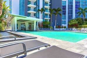 A hotel pool area with lounge chairs, a covered seating area, and a building with multiple balconies. - Modern 2-Bedroom Oasis with Pool (Surfers Paradise)
