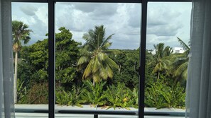 A view through a window with a view of a tropical landscape, including palm trees and banana plants. - Serene 2-bedroom Apartment With Mountain Views in Santiago (Santiago de los Caballeros)