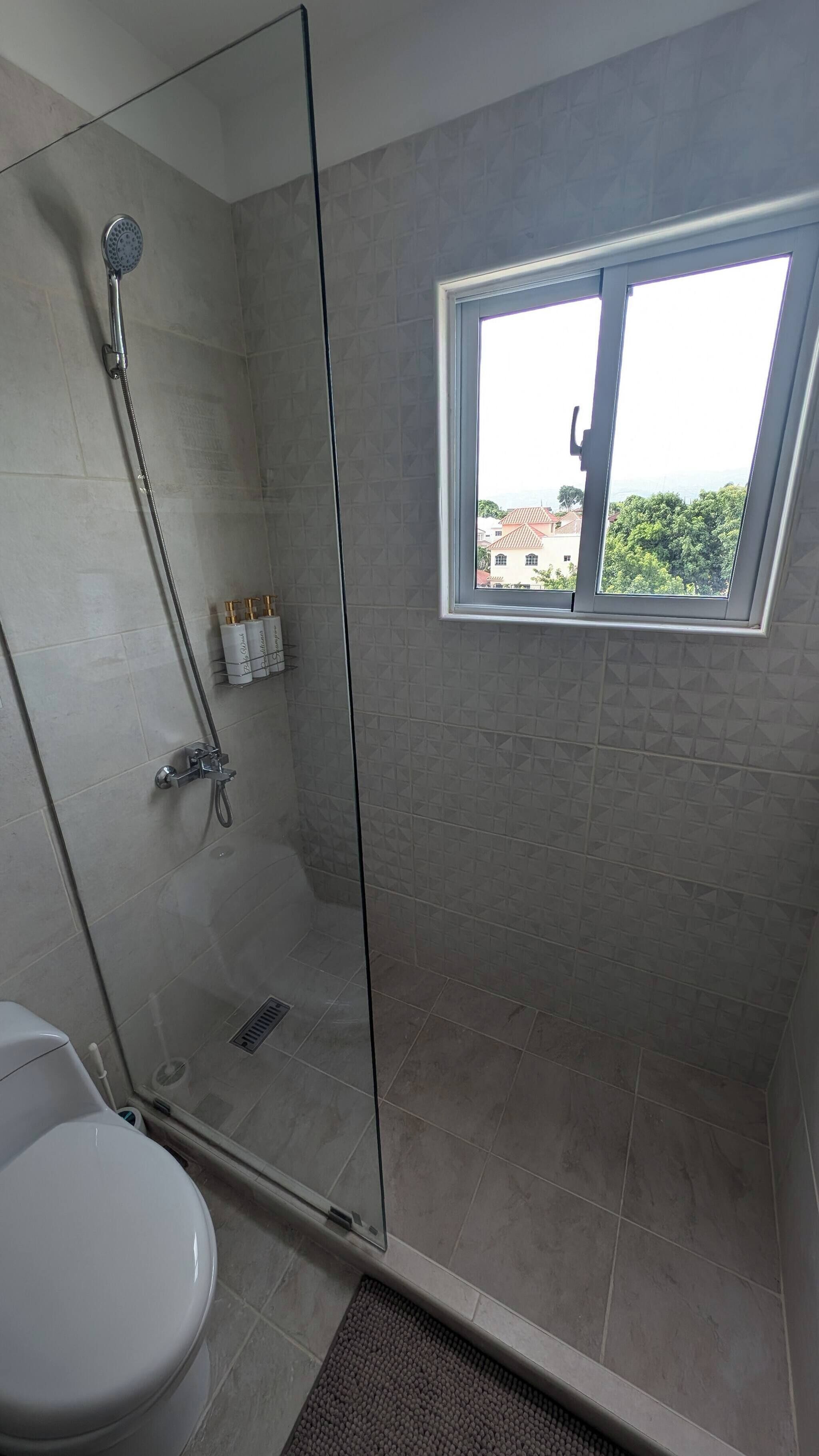 Un baño moderno con cabina de ducha de vidrio, inodoro y ventana con vista a edificios.