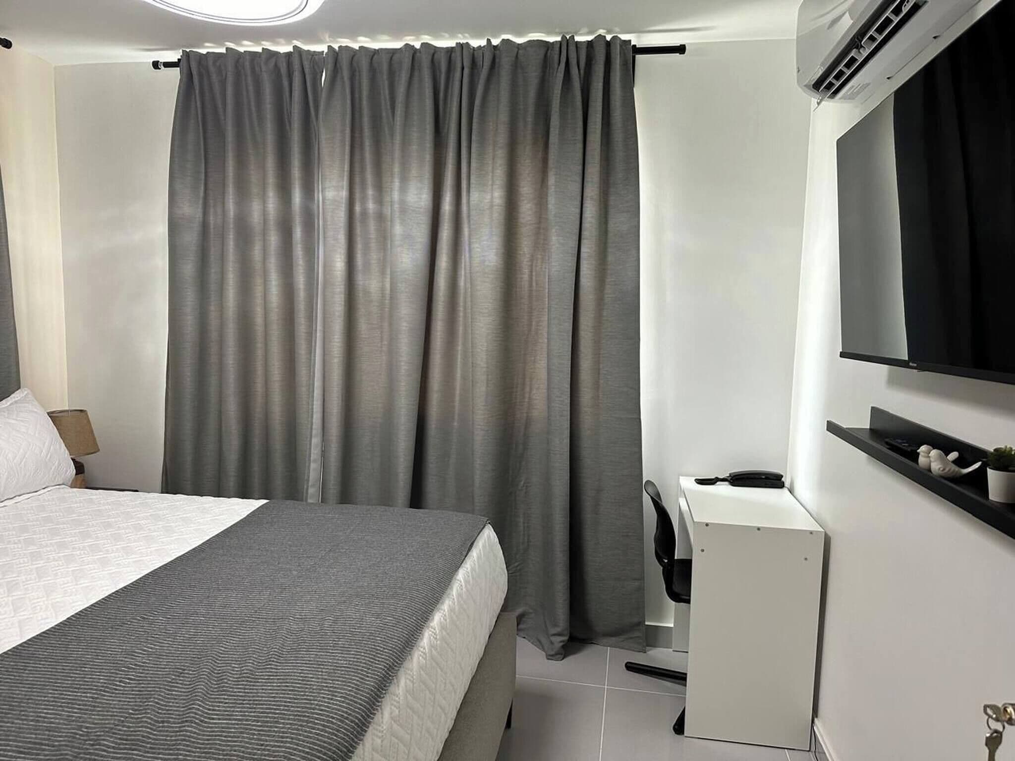 Ein Schlafzimmer mit Bett, Schreibtisch, Stuhl, Fernseher und Vorhang.