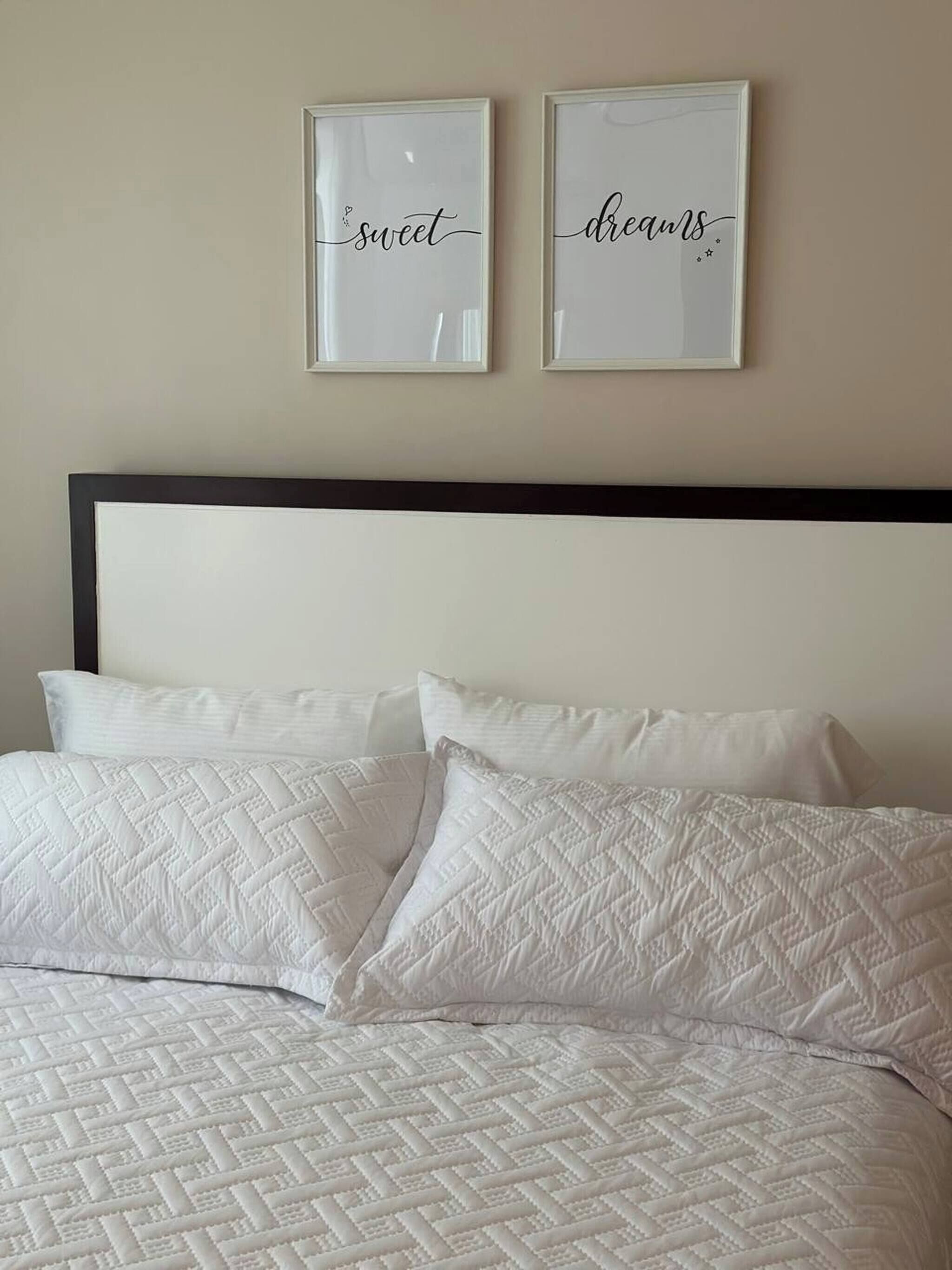 Un lit avec une couette blanche matelassée et deux oreillers, une tête de lit sombre et deux œuvres d’art encadrées accrochées au mur, comportant du texte.