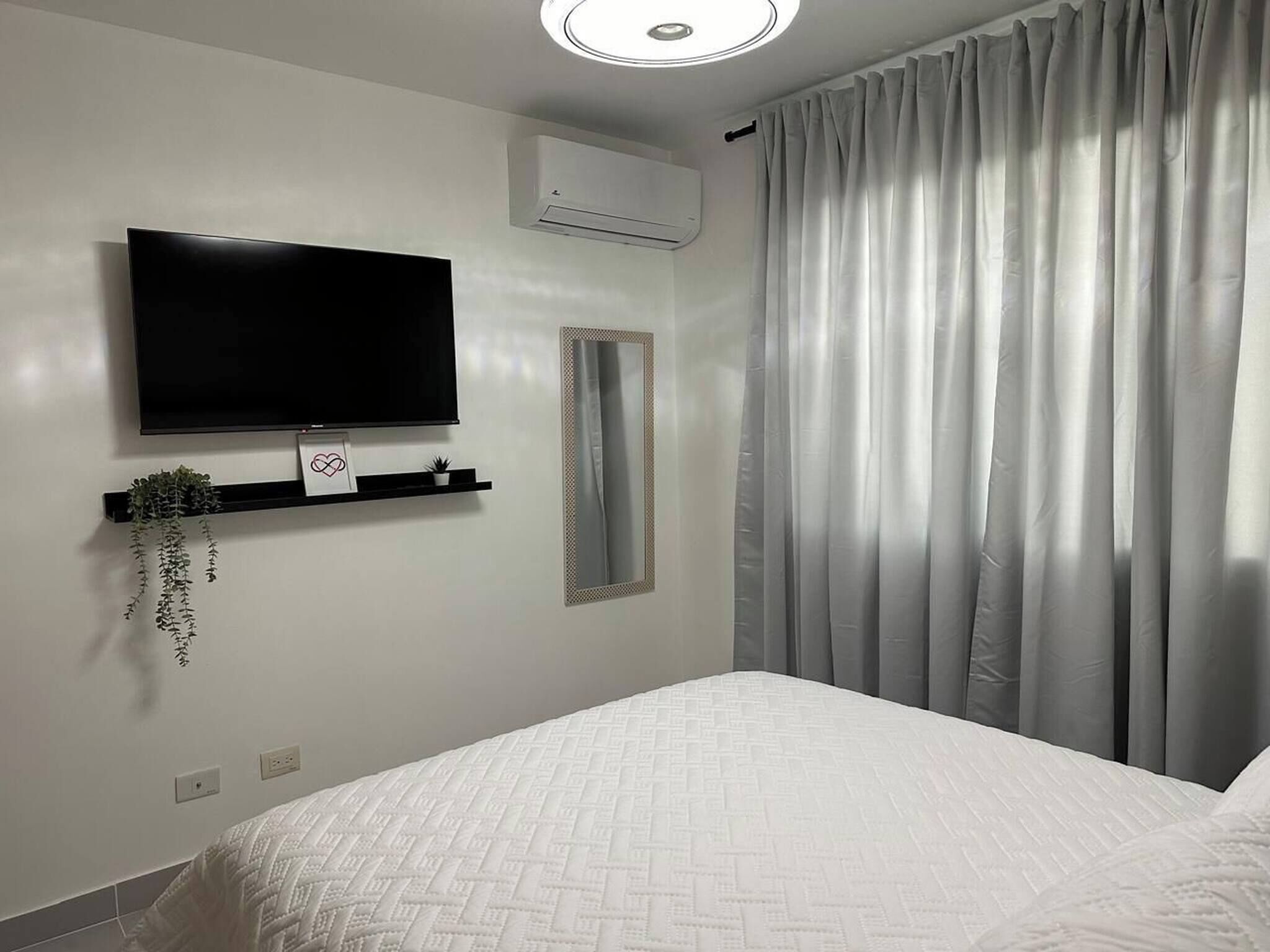 Un dormitorio con cama, televisor montado, aire acondicionado instalado en la pared y ventana con cortinas.