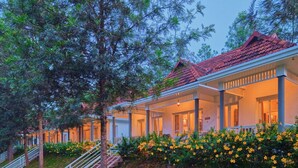 A row of white buildings with red roofs, connected by covered walkways, surrounded by greenery and yellow flowers. - Mistaura Home By Voye Homes (Udumbanchola)