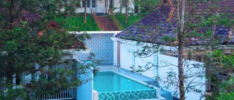 A swimming pool with a tiled deck, surrounded by greenery and a staircase leading to a building.