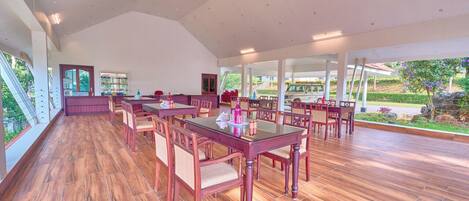 A spacious indoor area with wooden flooring, multiple tables and chairs, and large windows allowing natural light.