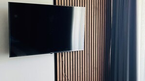 Una sala de estar moderna con un televisor de pantalla plana montado en la pared, un panel divisor de listones de madera, una planta en maceta y una cortina oscura.